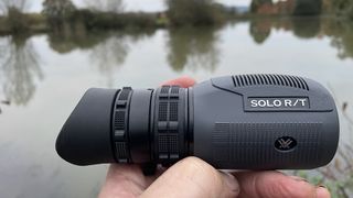 A close up of hand holding Vortex Solo R/T 8x36 monocular in front of a lake.