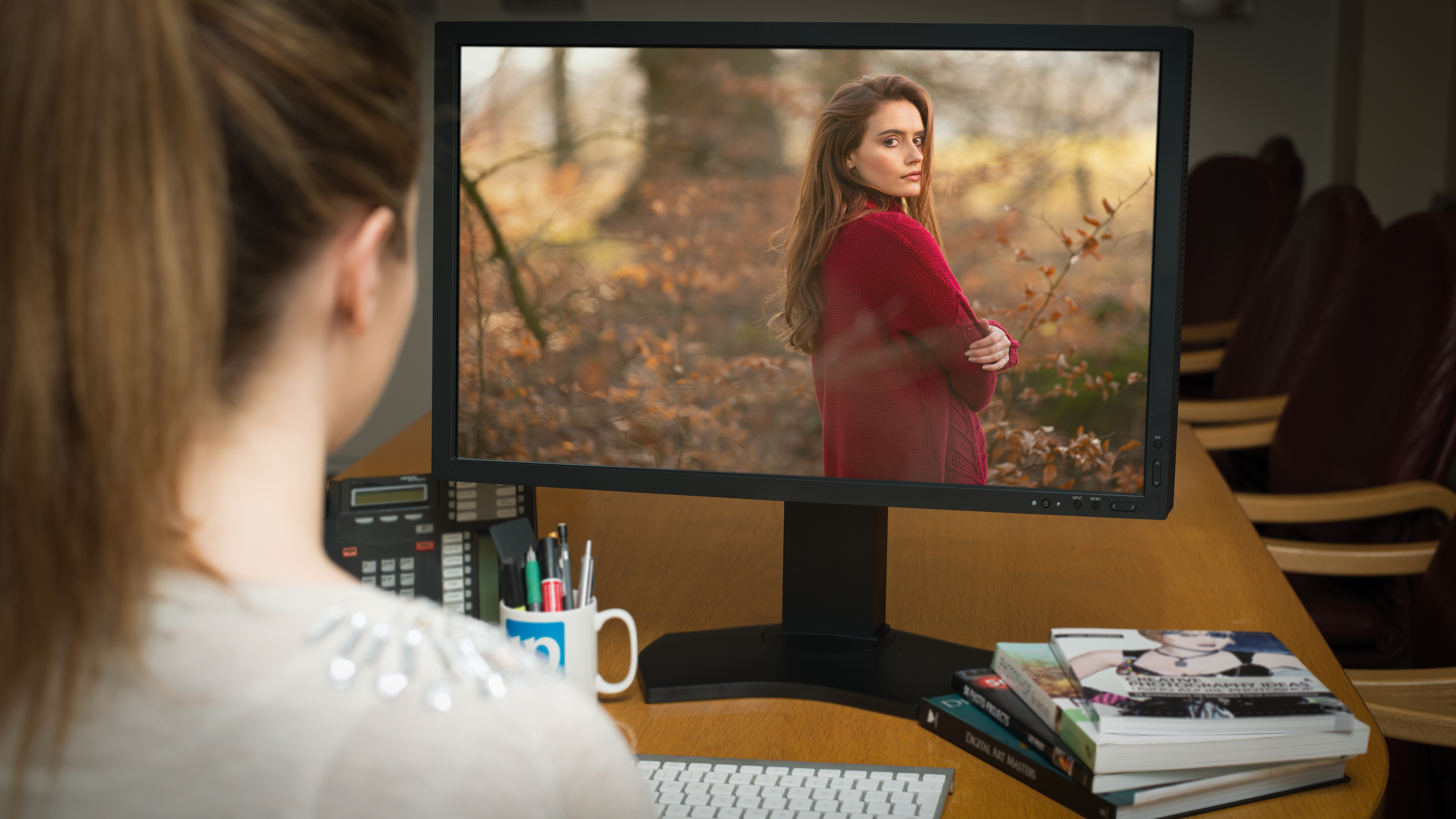 A person with a ponytail sitting at a computer, editing a portrait photograph of a young woman in a forest