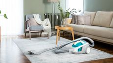 A blue and white vacuum cleaner on a cream living room rug, beside a cream couch and a small yellow side table