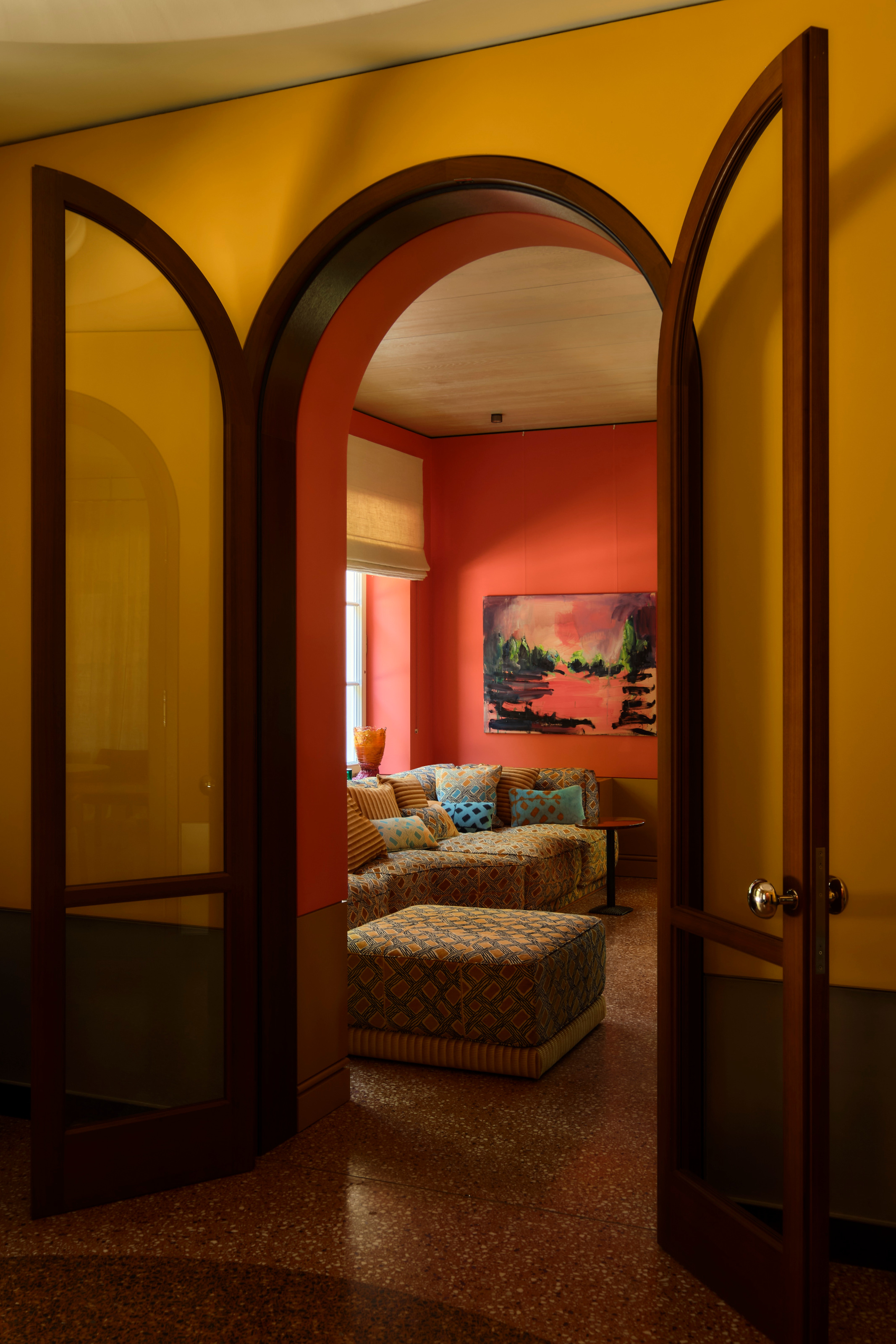 View from ochre hallway through arched glazed doors into snug with warm red walls and blue and yellow patterned sofa and footrest