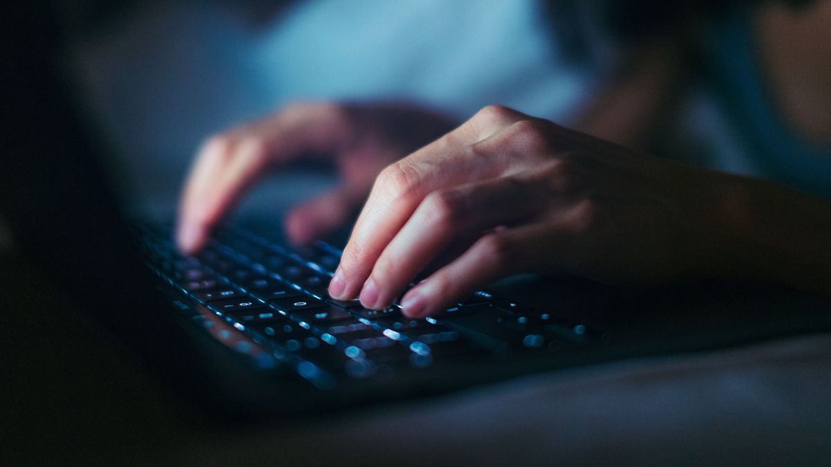 A close-up photograph of a person&#039;s hands typing on a backlit laptop keyboard