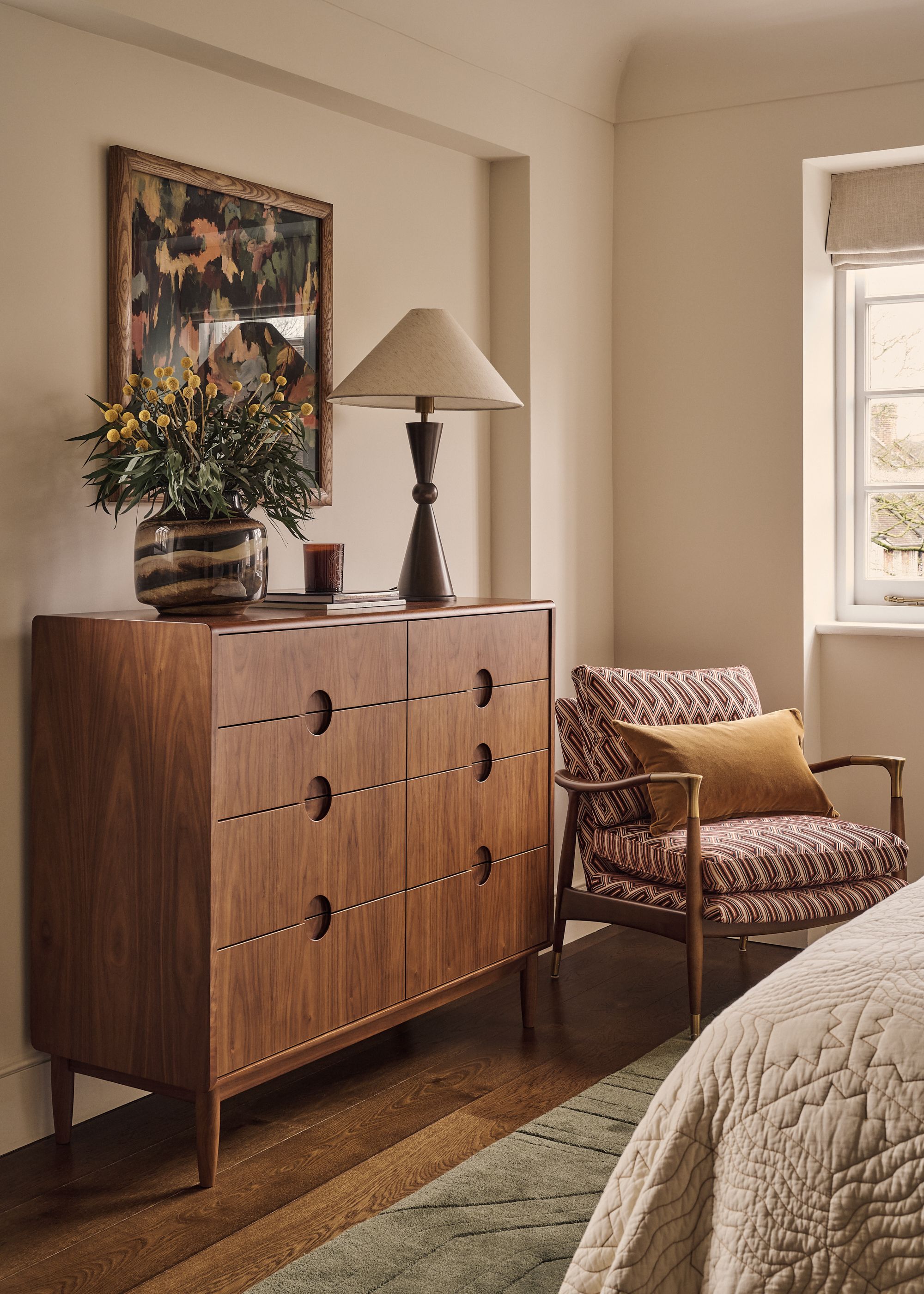 A modern bedroom with a framed print, a vase of flowers, a table lamp, a wooden chest of drawers, a wood armchair with patterned cushions, and a bed