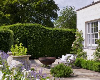 A sunny paved garden with a firepit, bench with white cushions and white exterior wall with windows