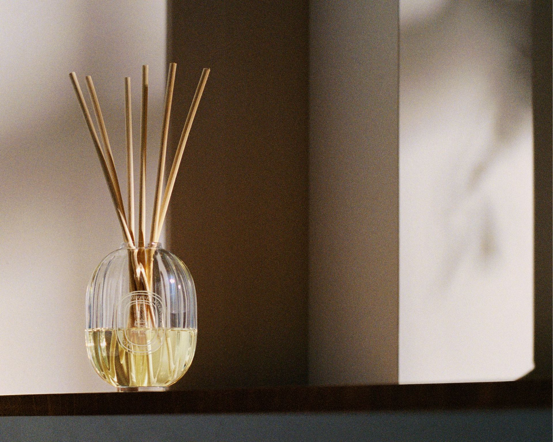 A glass Diptyque diffuser with light wooden reeds against a shadowy wall
