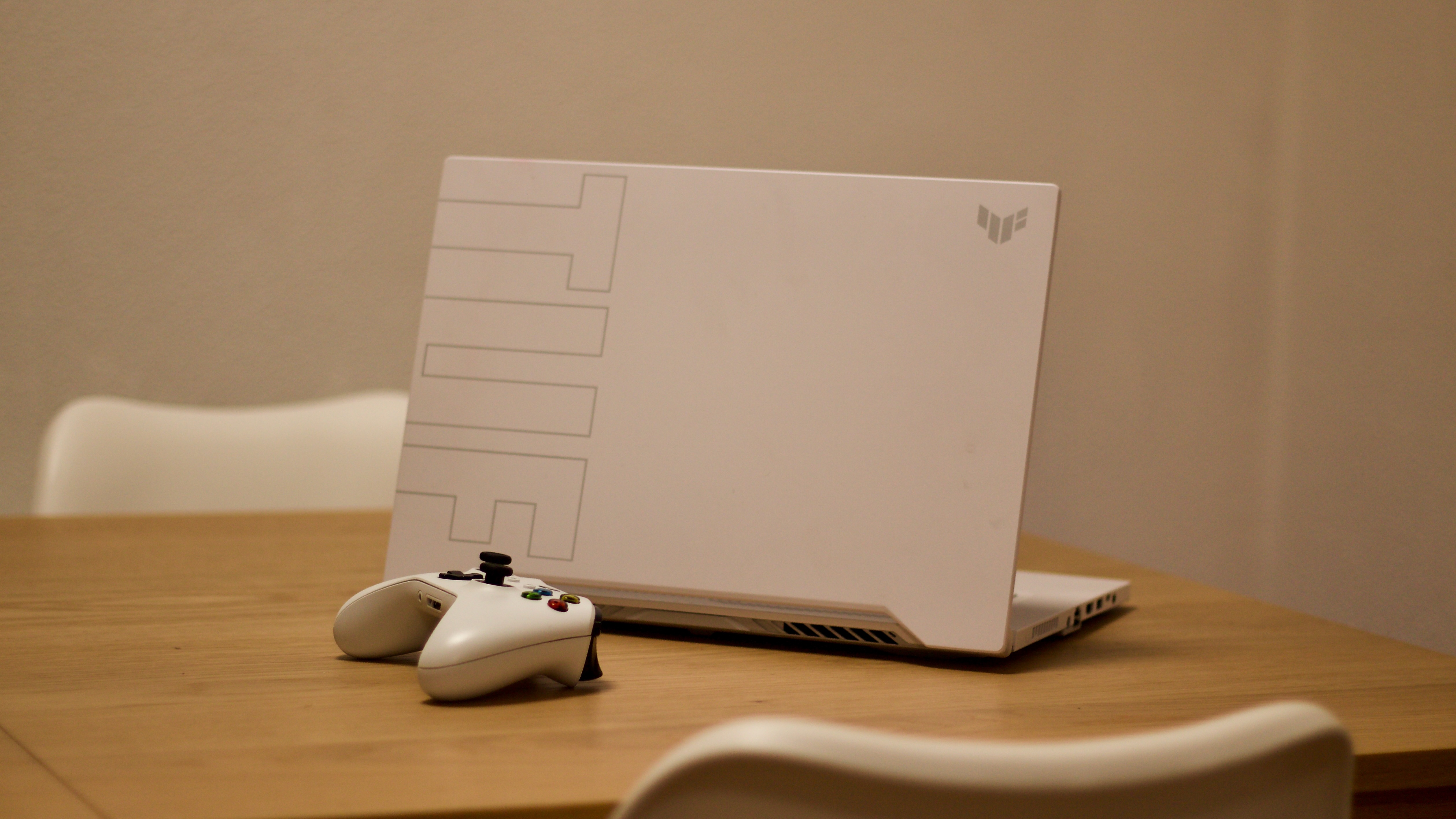 En hvit ASUS TUF Dash F15 står åpnet på et trefarget bord, med en hvit Xbox-spillkontroller liggende ved siden av.