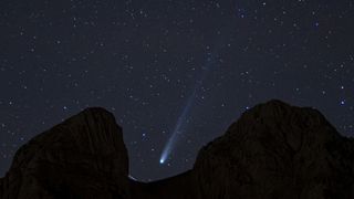 A white streak of a comet's tail can be seen in the center of the image as it streaks diagonally from top right to lower left between the dark silhouette of rocks blocking part of the starry night sky
