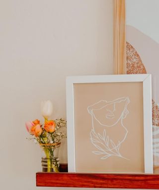 A brown wall art print of a face outline on a wall shelf next to a vase of orange roses and pink white tulips