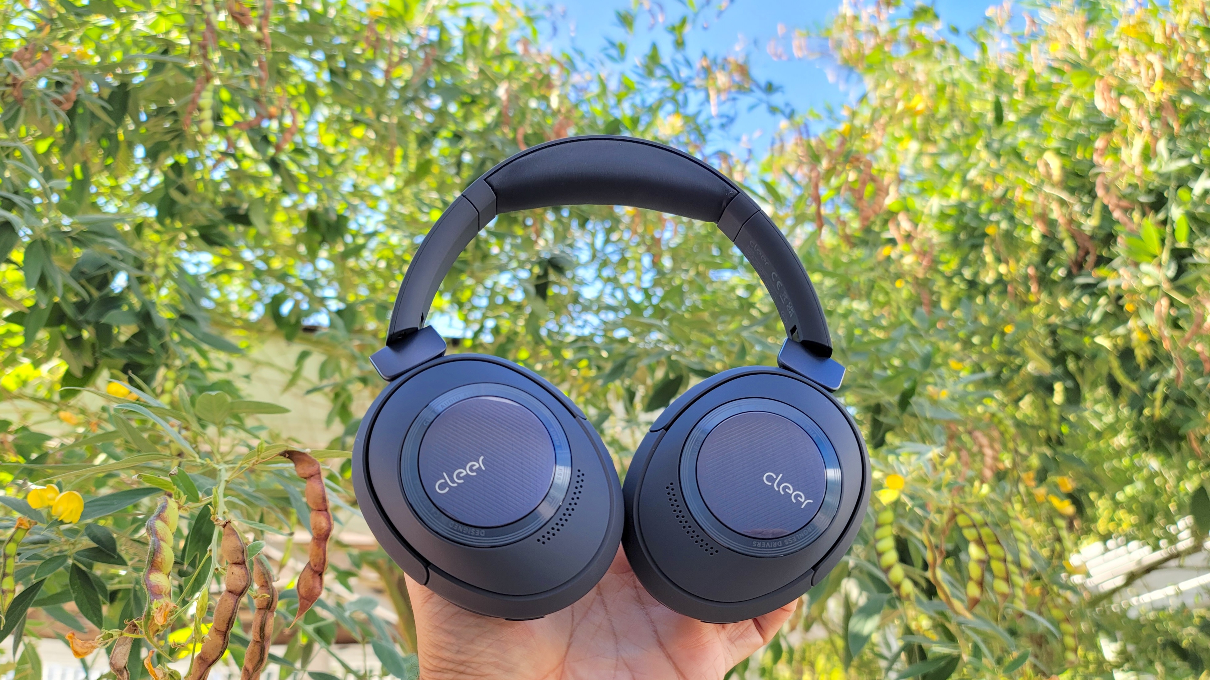 The Cleer Audio Alpha headphones being helf aloft a backdrop of trees