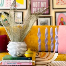 Vase with faux leafy plant sitting on pile of coffee table books beside candle holder with four lilac candles, in front of mustard sofa and colourful gallery wall