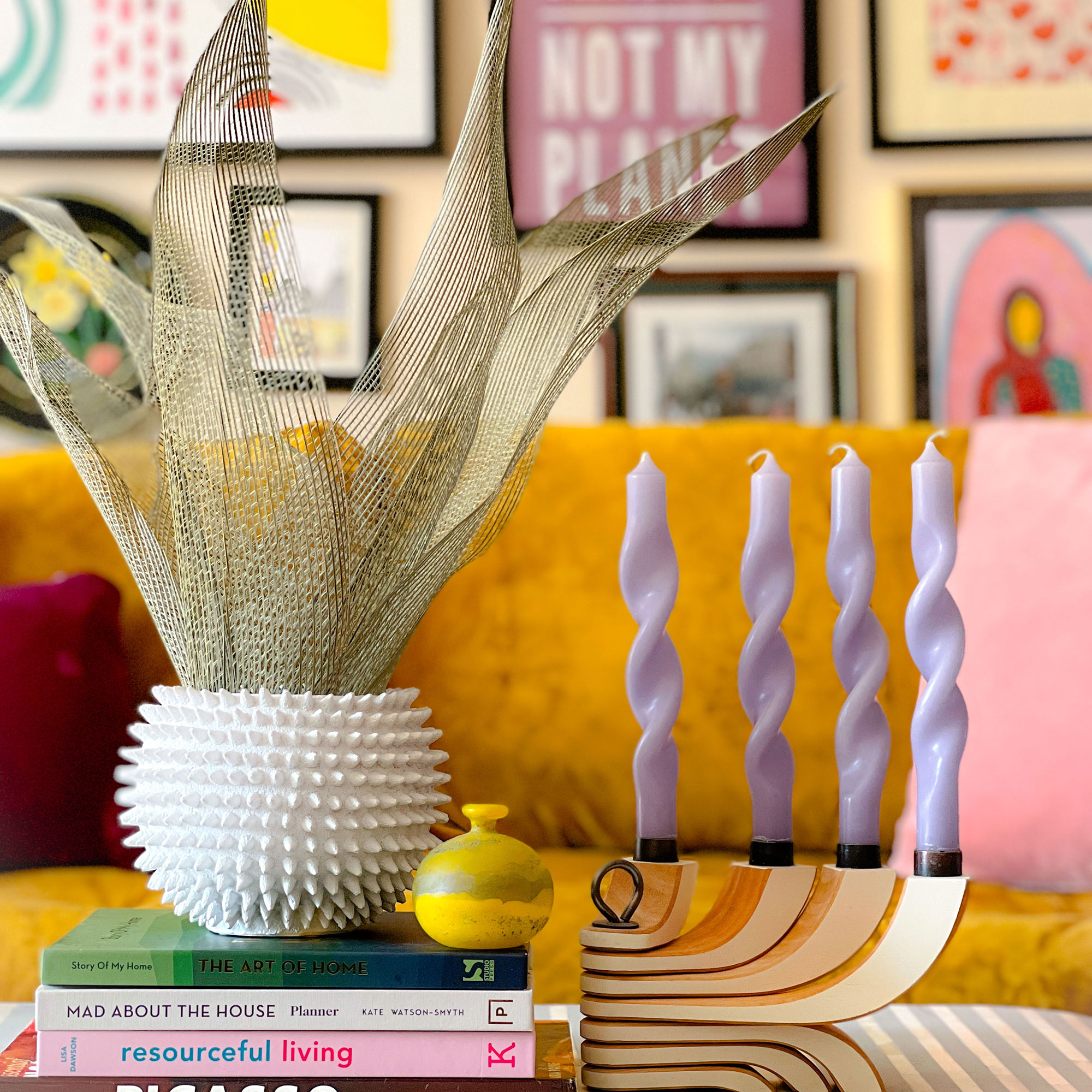 Vase with faux leafy plant sitting on pile of coffee table books beside candle holder with four lilac candles, in front of mustard sofa and colourful gallery wall