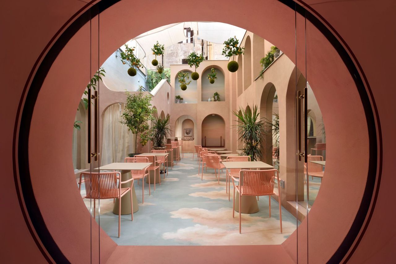 A surreal hotel&#039;s inner courtyard features rose and blue-tinted décor, including sky-like floors, pink curtained vaults, and trailing plants. 