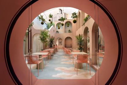 A surreal hotel's inner courtyard features rose and blue-tinted décor, including sky-like floors, pink curtained vaults, and trailing plants. 