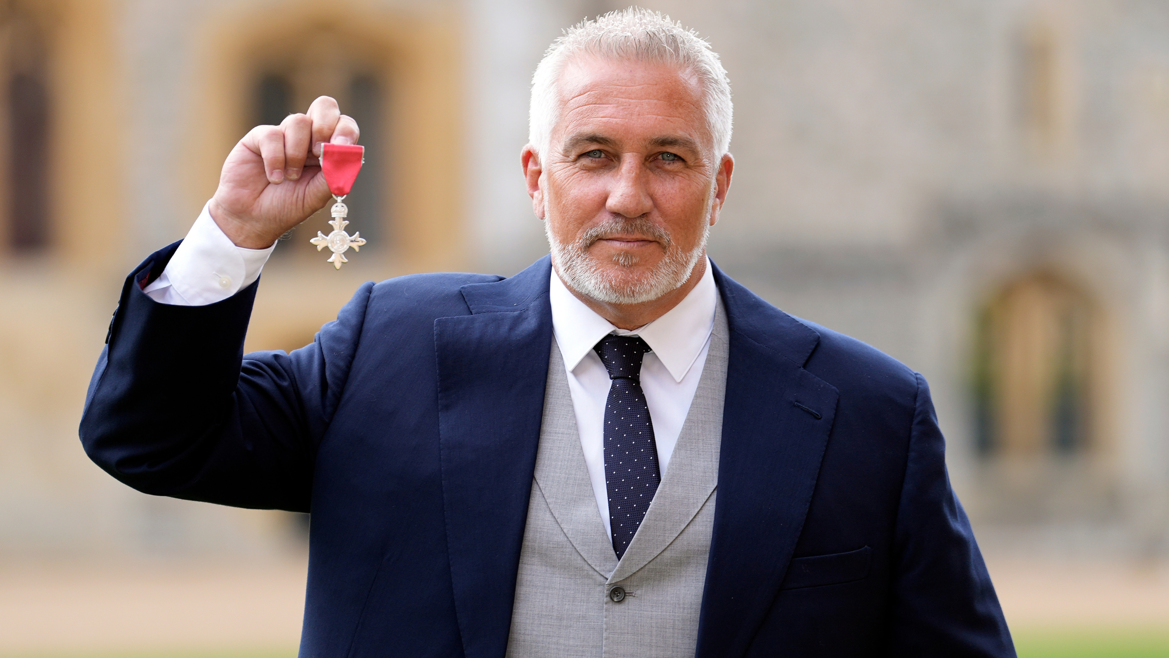 Paul Hollywood in navy suit after being made a Member of the Order of the British Empire. He is holding up his medal.