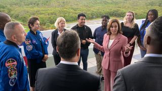 a woman in a salmon-colored suit talks to a crowd of people that includes several astronauts in blue flight suits.