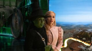 Cynthia Erivo and Ariana Grande stand smiling next to a model of Oz in Wicked.
