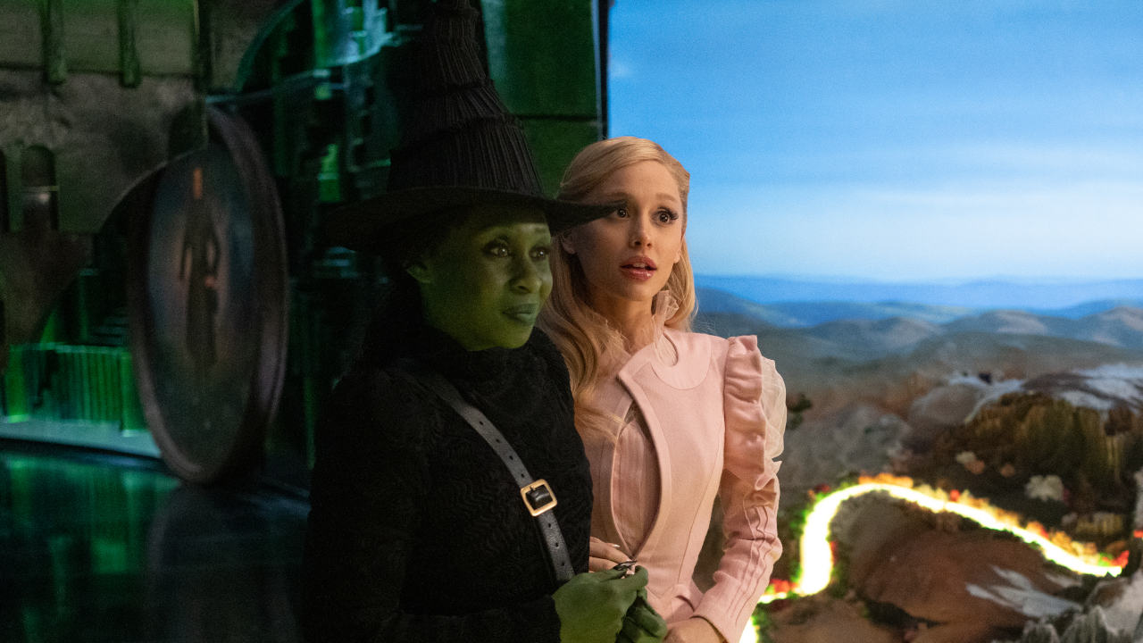 Cynthia Erivo and Ariana Grande stand smiling next to a model of Oz in Wicked.