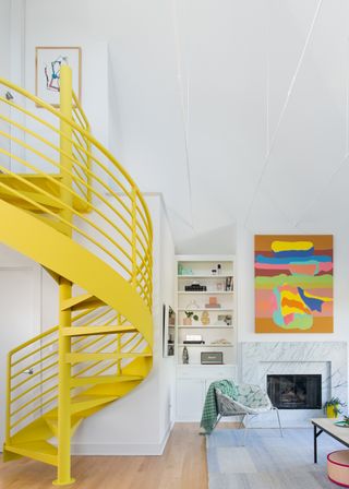 white room with bright yellow spiral staircase leading down to the living room with marble fireplace, colorful art hanging above and styled shelves