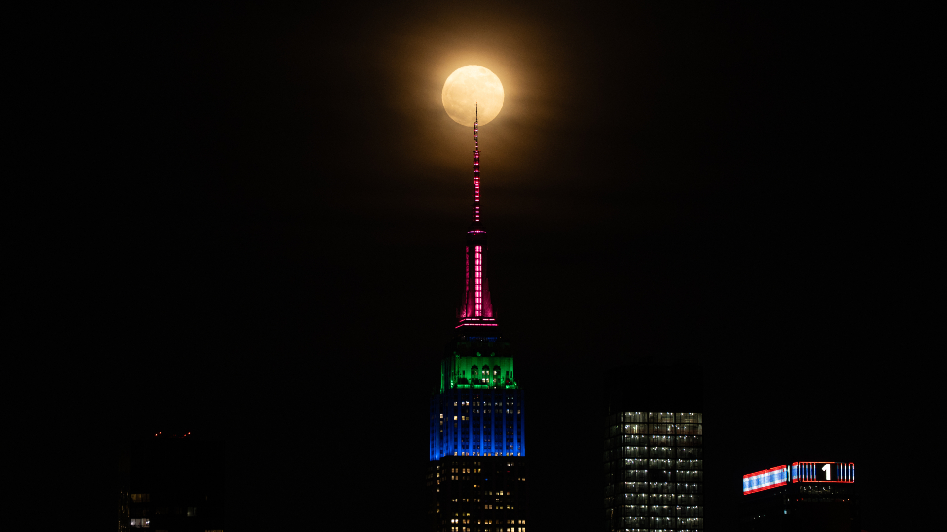 The full yellow hued disk of the flower moon is pictured behind the spire of the Empire State Building in New York City, hanging against a dark sky. The tops of two other skyscrapers can be seen to the lower right of the image.