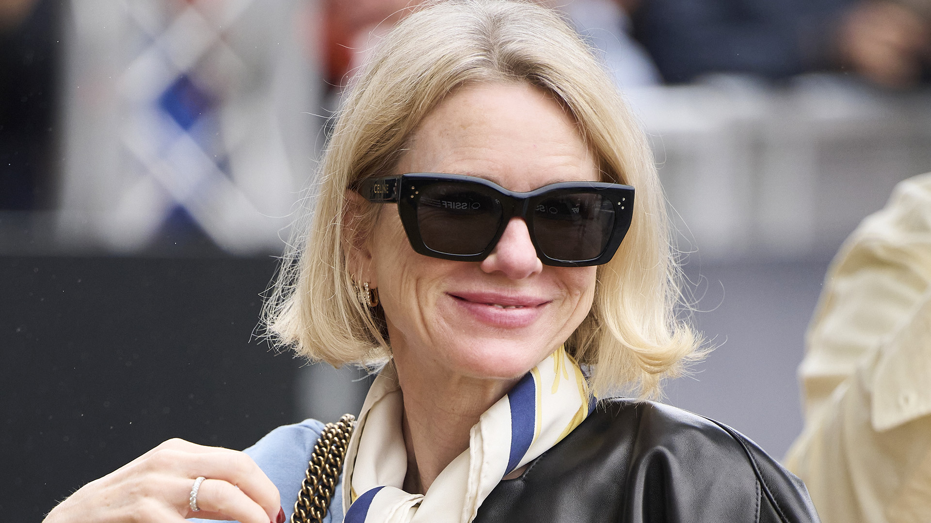 Naomi Watts in in San Sebastian, Spain wearing a leather bomber jacket, neck scarf, light blue sweater, bootcut jeans, and Adidas Samba sneakers