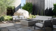 An outdoor minimalist garden space with white furniture, a fire pit, a large rock, and textured plants