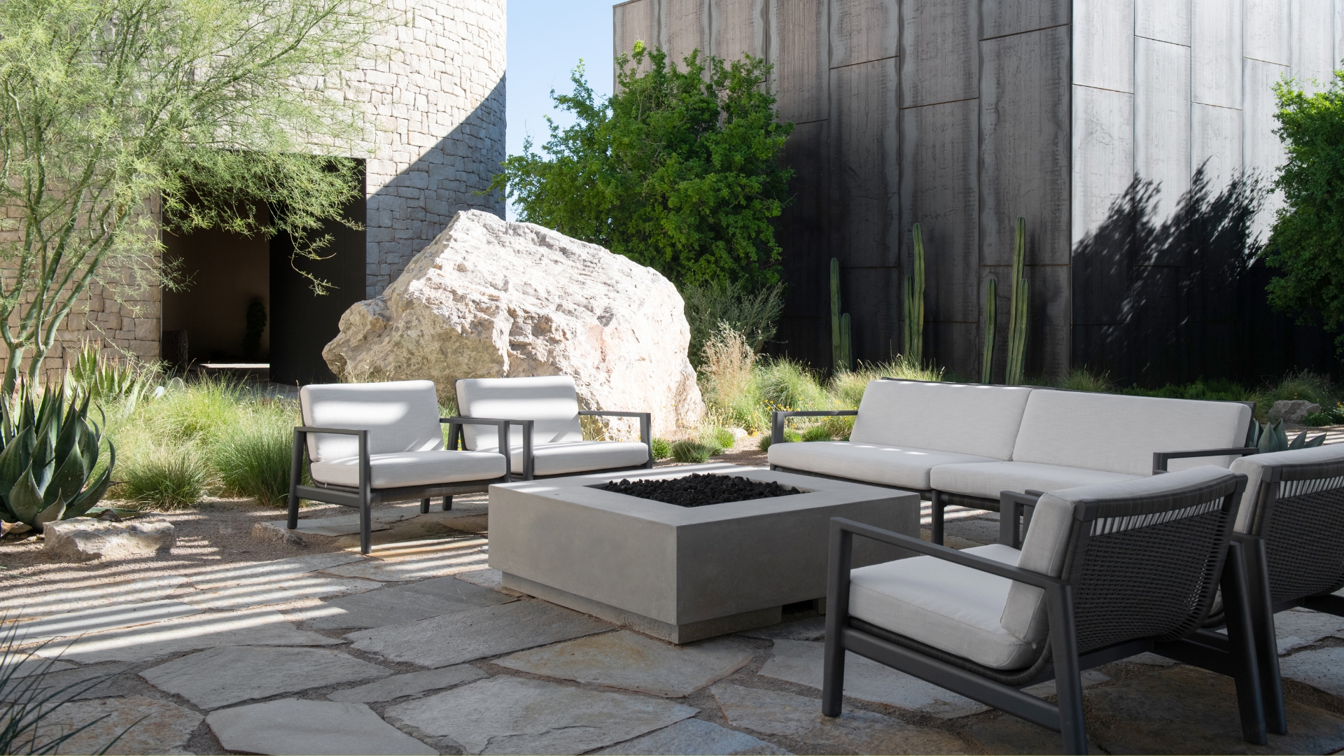 An outdoor minimalist garden space with white furniture, a fire pit, a large rock, and textured plants