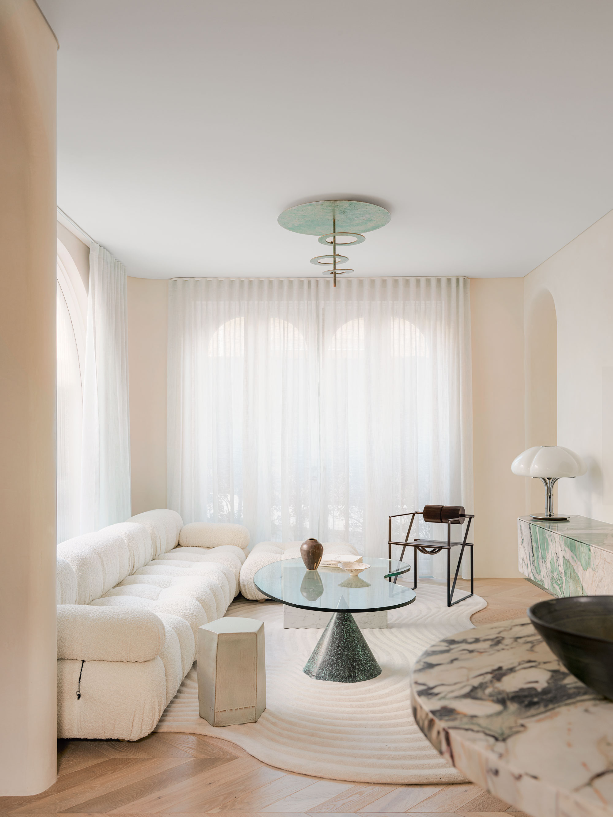 A cream colour drenched living space with a large cream sofa to the right and a marble table to the right, a glass coffee table and floor to ceiling, sash curtains.
