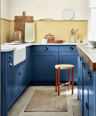 cobalt blue kitchen with yellow painted backsplash tiles, marble countertops, butler sink, stool, wooden chopping boards, rug, artisan ceramics, white wall above