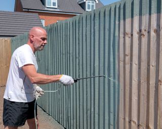 Man using a paint sprayer to paint a garden fence green