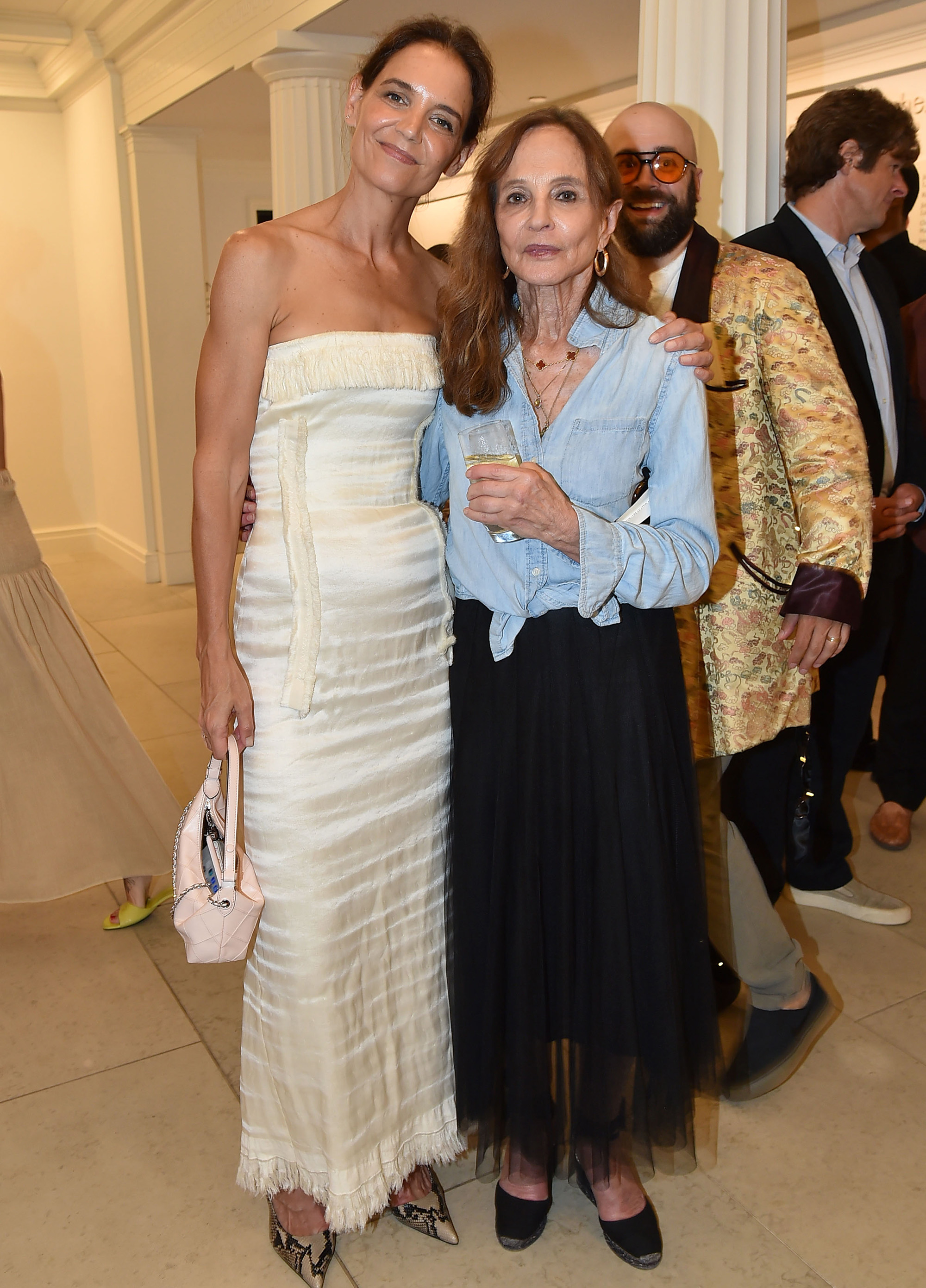 Katie Holmes getting photobombed from behind in yellow cream dress at a Hamptons Gala summer 2026.