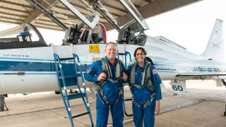 two astronauts in flight suits in front of an airplane with two cockpit hatches open
