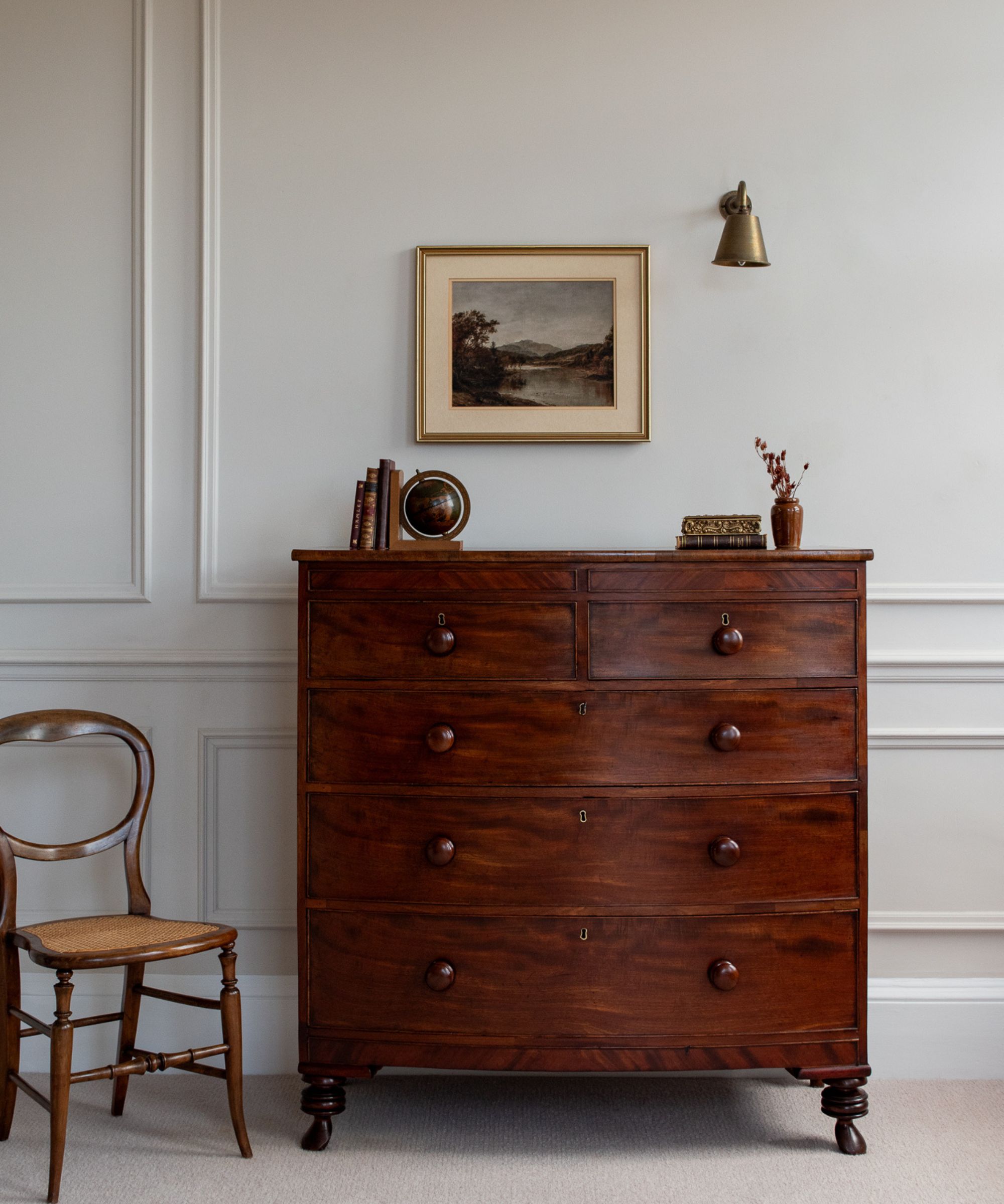 White room with a dark mahogany chest of drawers at the centre, antique decor on top, a landscape painting above it, a gold sconce above that, and an antique cane chair to the left.