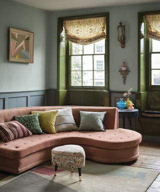 living room with two tone blue walls, green painted windows, pink curvy couch, rug, floorboards, pink, green and blue couch pillows, side table, artwork