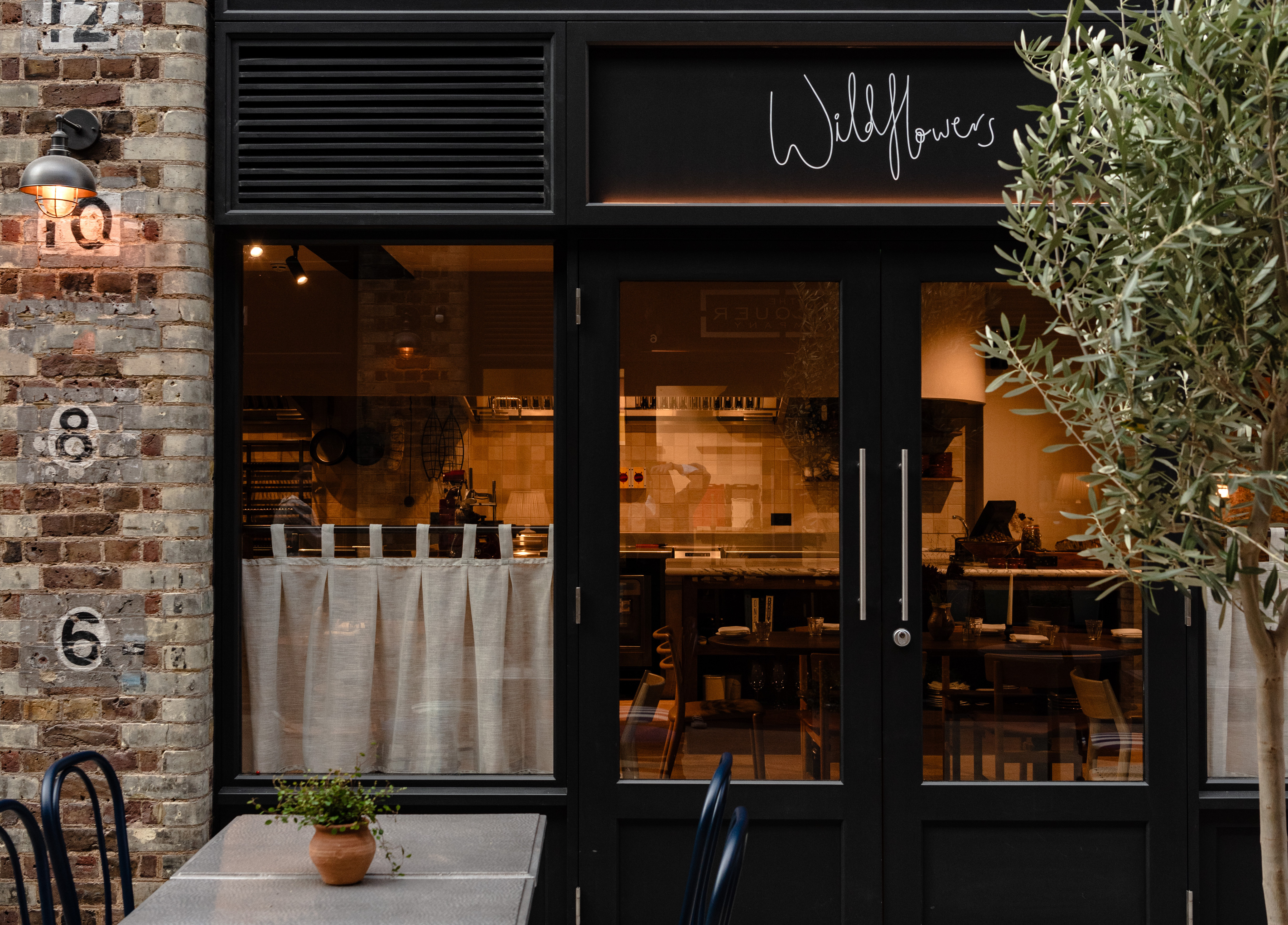 A brick restaurant exterior, decorated with olive trees, a black-painted entrance with the sign "wildloflowers" in white handwriting, and a warm atmosphere inside.