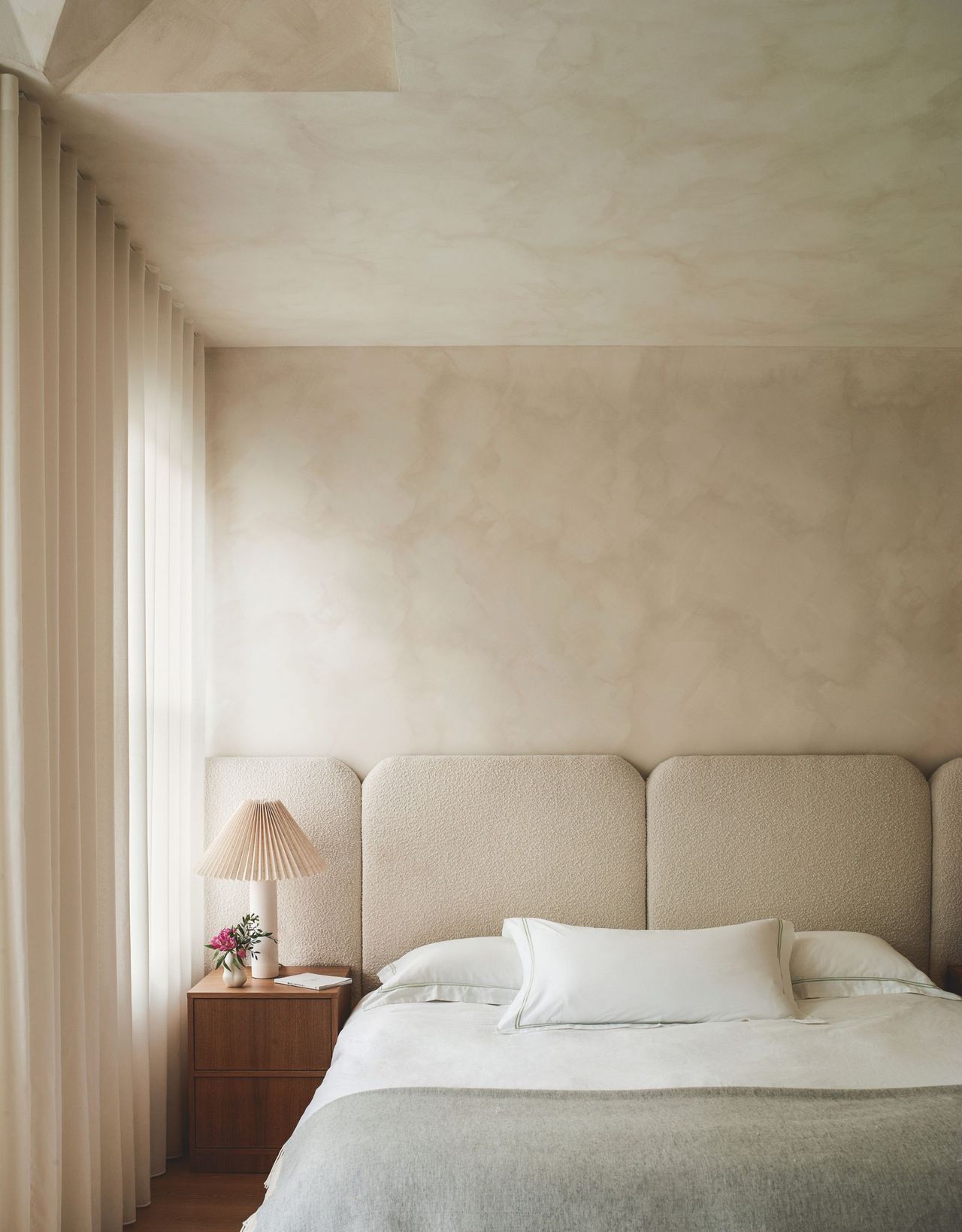 A modern neutral bedroom with an extended headboard right to the end of the wall and sheer curtains and limewashed walls