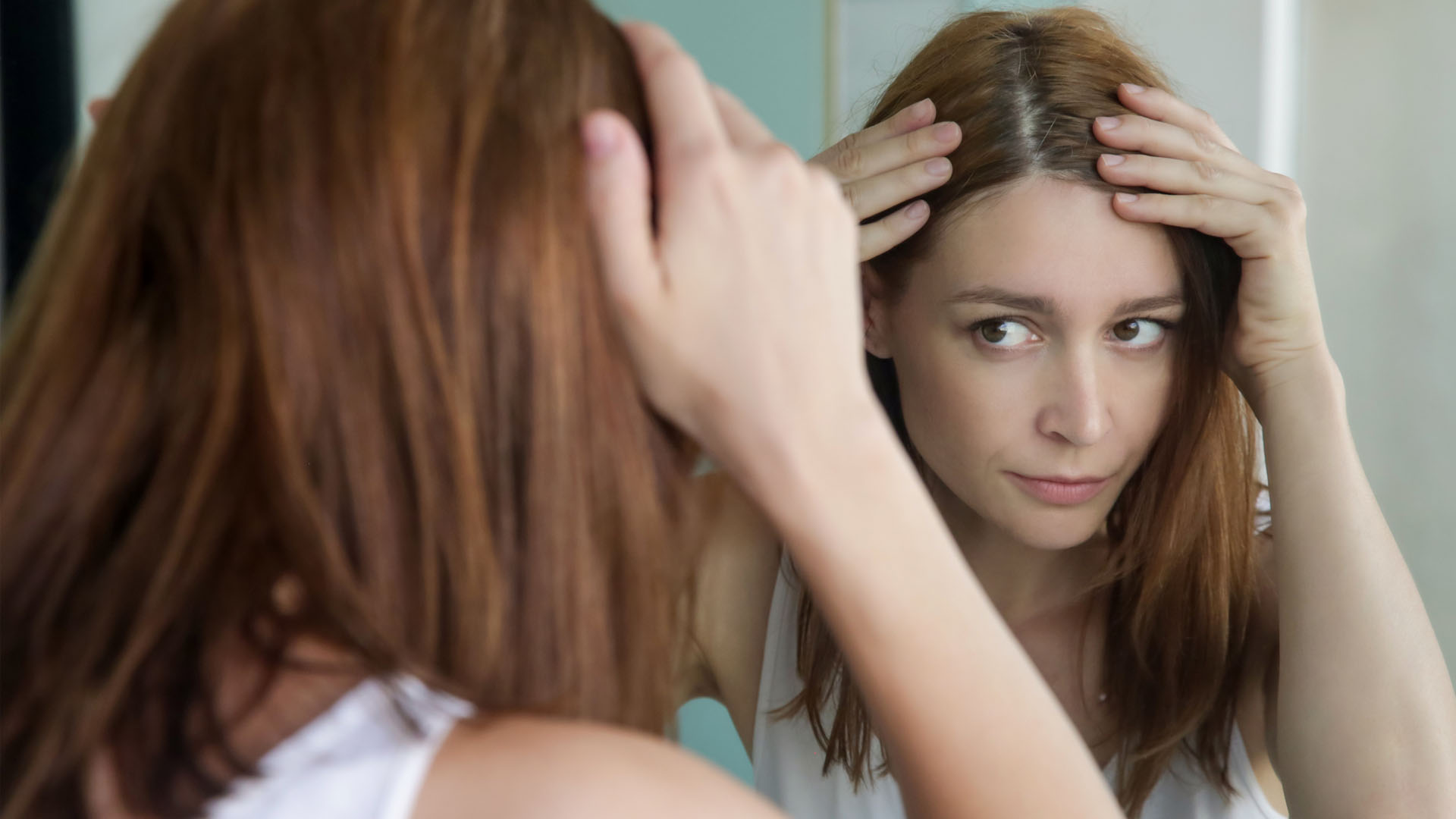 Image depicts a woman inspecting her hair for grays in a mirror