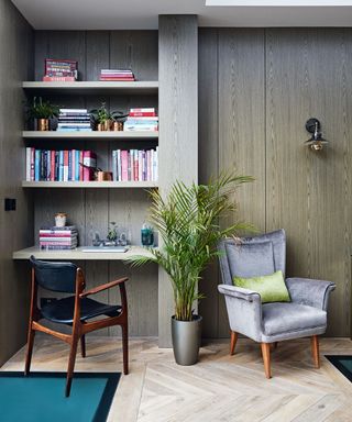 Corner of a living space with built in desk and shelving for books, brown-gray wooden walls, dark wood and black leather desk chair, upholstered gray armchair with green cushion and wooden legs, metal wall light above armchair, green grassy plant in gray plant pot