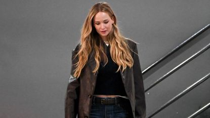 Jennifer Lawrence wearing blue jeans with a brown leather blazer, a black top, and a black handbag