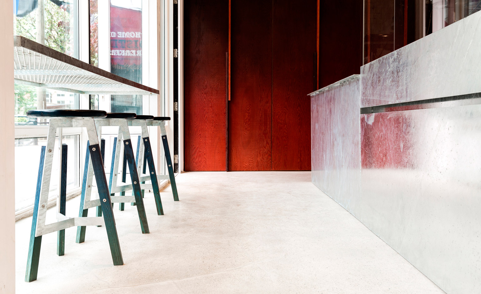 Wood and metal based stools under a mesh counter infront of a large glass window in a room with red wood paneling and a grey counter