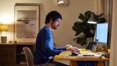 A remote worker sat at his desk, in a plush modern apartment lit by lamps.