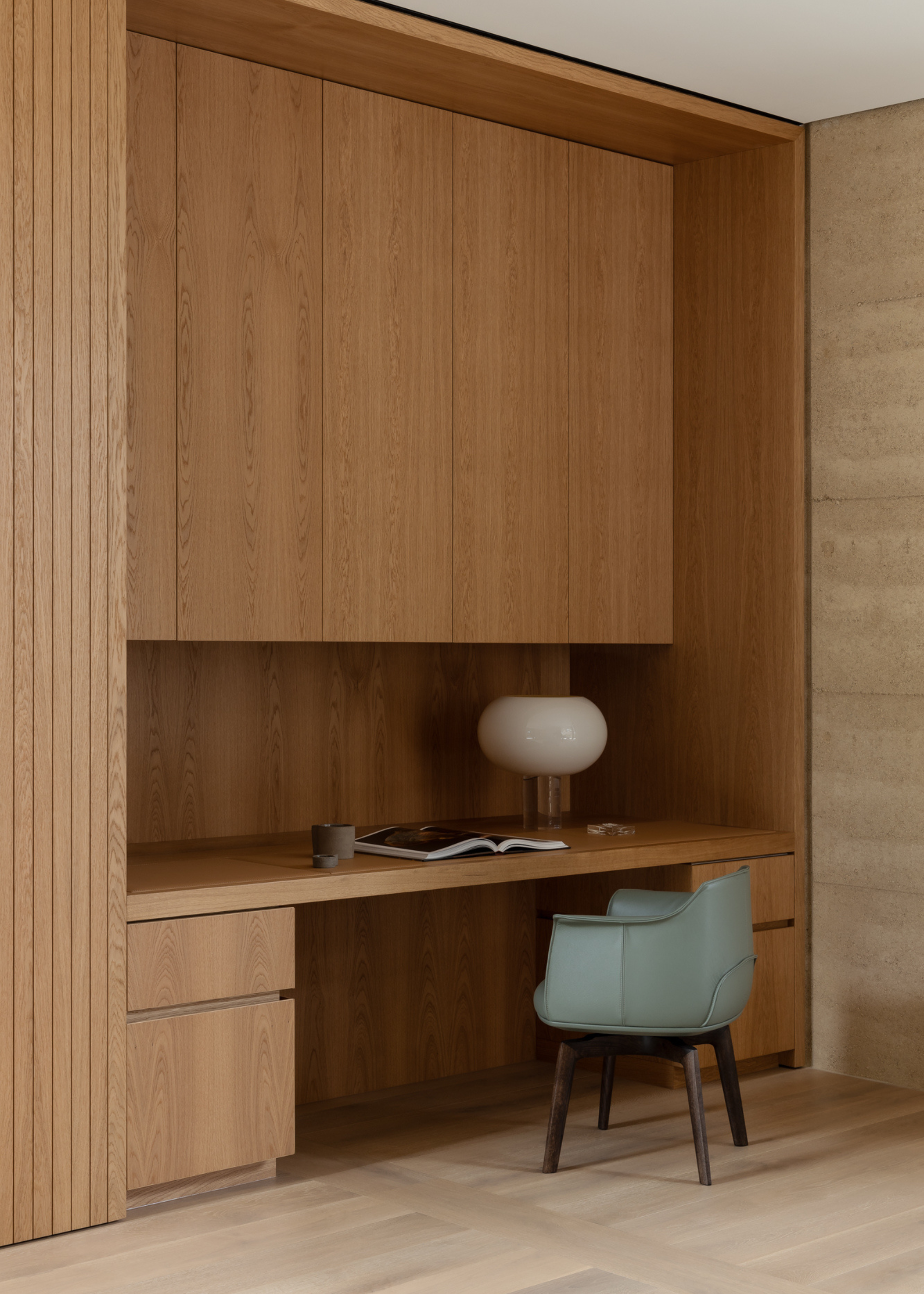 A minimalist home office with a wood desk, wood storage cabinets, a blue armchair, and a table lamp beside a ceramic cup and an open book