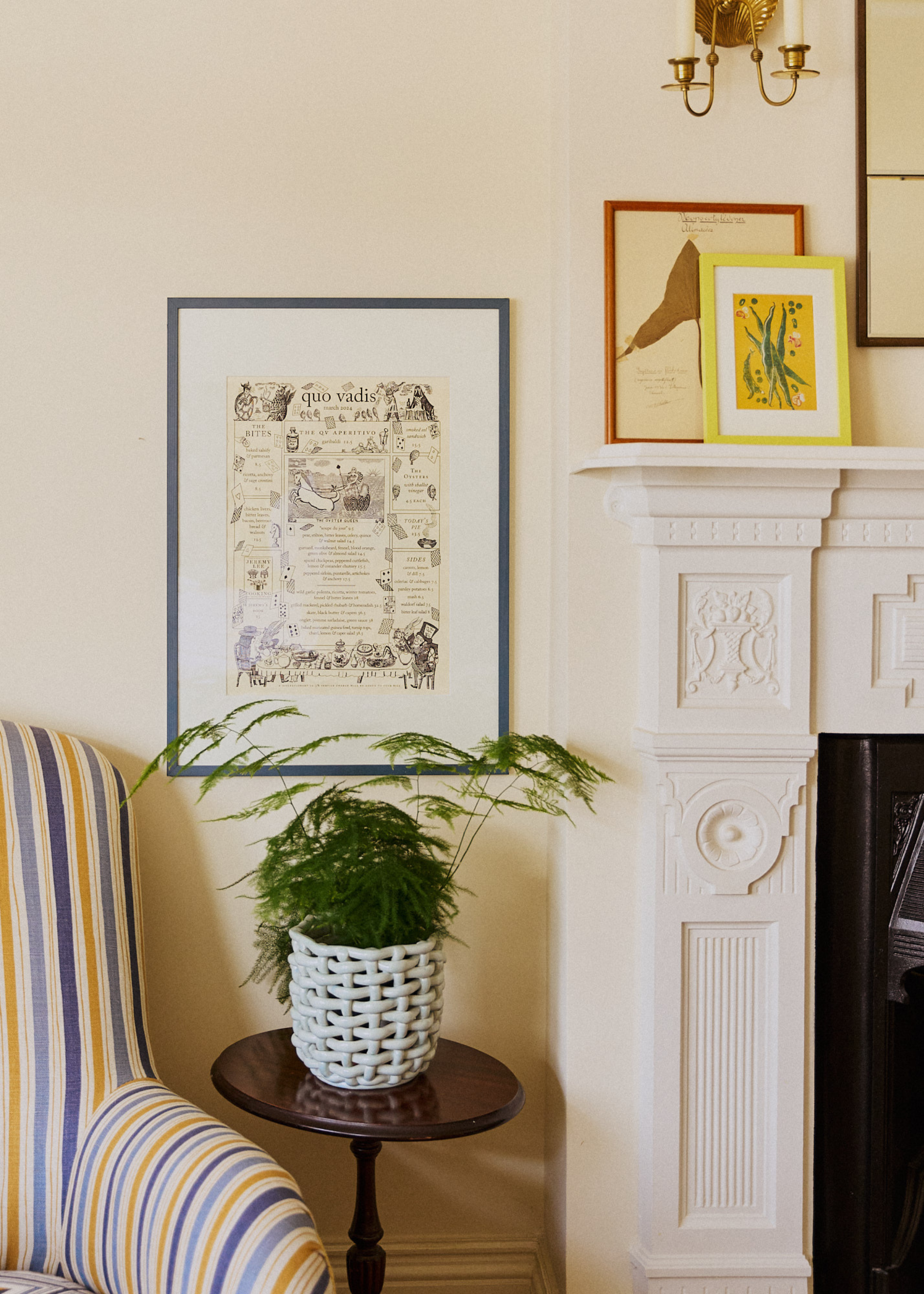 A living room with a striped yellow and blue sofa seat chair beside a wood pedestal with a basket-weave planter with a fern. A framed print on the wall and a pair on the mantel above the fireplace
