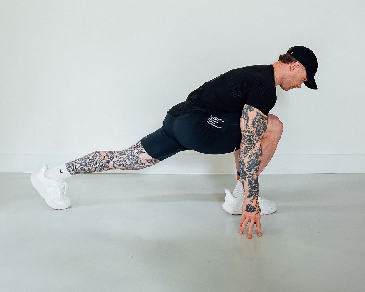 Coach Corey Perkins performs a low lunge. His front leg is bent at a 90 degree angle and his back leg is stretched behind him. He holds his torso close to his front leg and has placed both hands on the floor for balance.