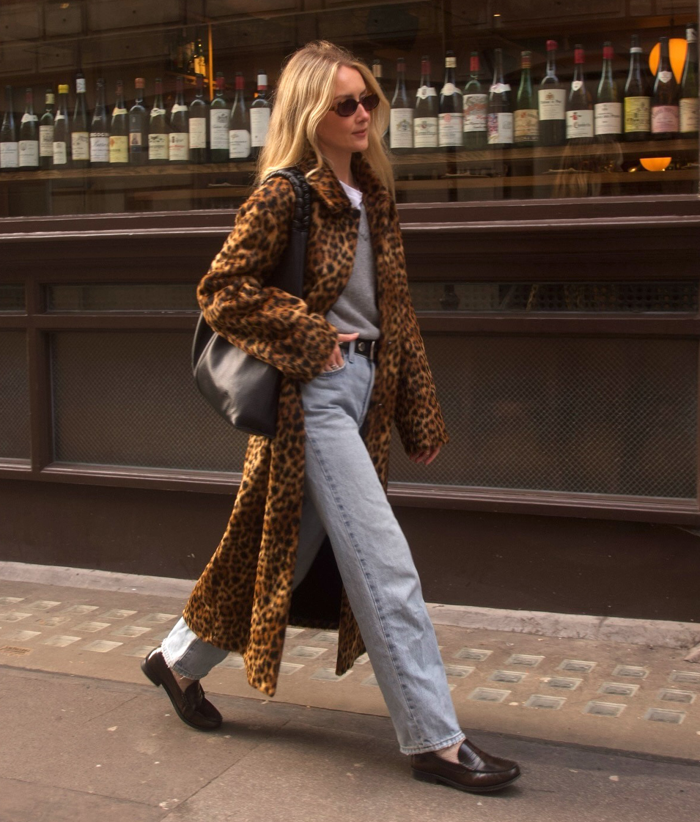 British style influencer Chloe Kath Butler walking on a London sidewalk wearing a leopard-print coat, gray V-neck sweater layered over a white T-shirt, studded black belt, black tote bag, light-wash jeans, and brown loafers