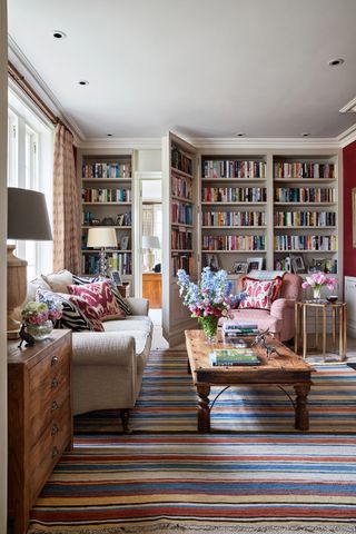 Living room with cream upholstered sofa and rustic wooden coffee table on striped rug bookcases with hidden door on far wall