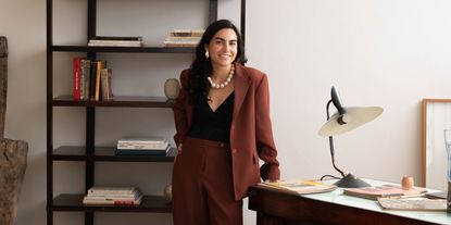 A young woman dressed in a terracotta brown loose suit, paired with a black, V-neck body and a chunky cream necklace and earrings stands smiling in a mid-century modern-style office.