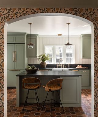 Sage green painted kitchen cabinets in a 1920s revival house, with white clapboard walls and hexagonal terracotta floor tiles.