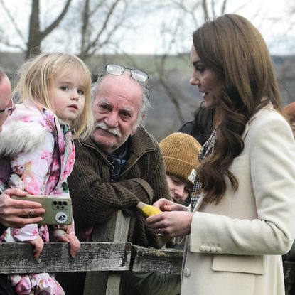 Kate Middleton wears a long cream coat with a dogtooth print dress and speaks to a young girl in the crowd