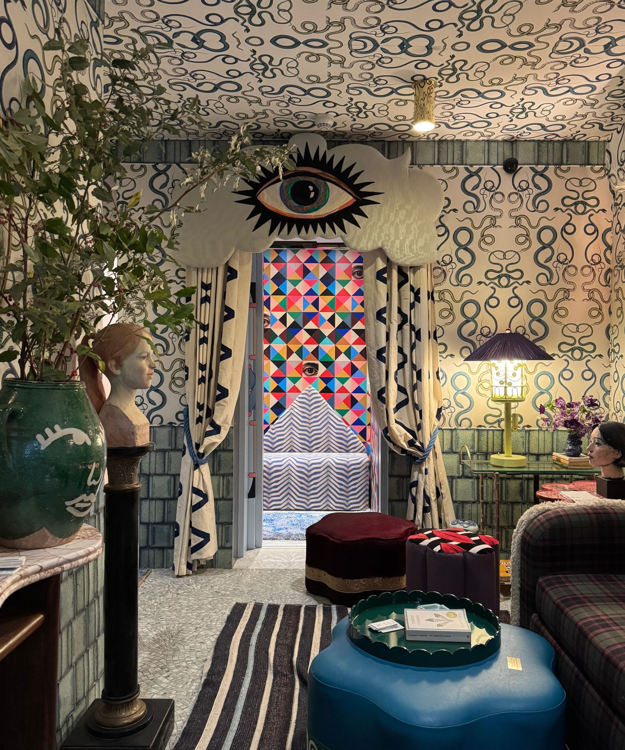 BK Showhouse living room with large eye pelmet over window looking to more eyes on multicolored wallpaper; maximalist wallpaper on walls and ceiling and footstools/sofas in rest of room