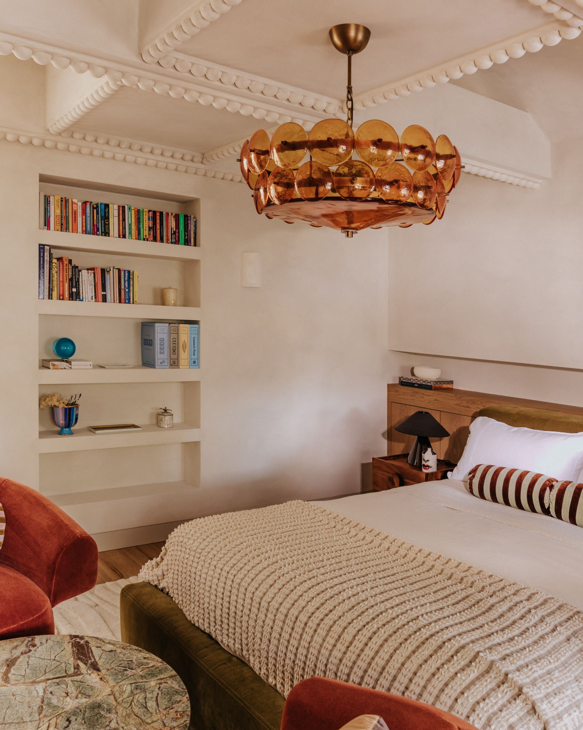 Image of a lime wash beige bedroom with built-in bookshelves, a bed with white and red and white stripped bedding. There is a amber glass pendant chandelier on the ceiling.