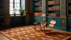 a study room with a large rug with pink zig zag stripes and a brown background, teal painted bookshelves holding many books, and a wooden chair and an accent table in the middle of the room